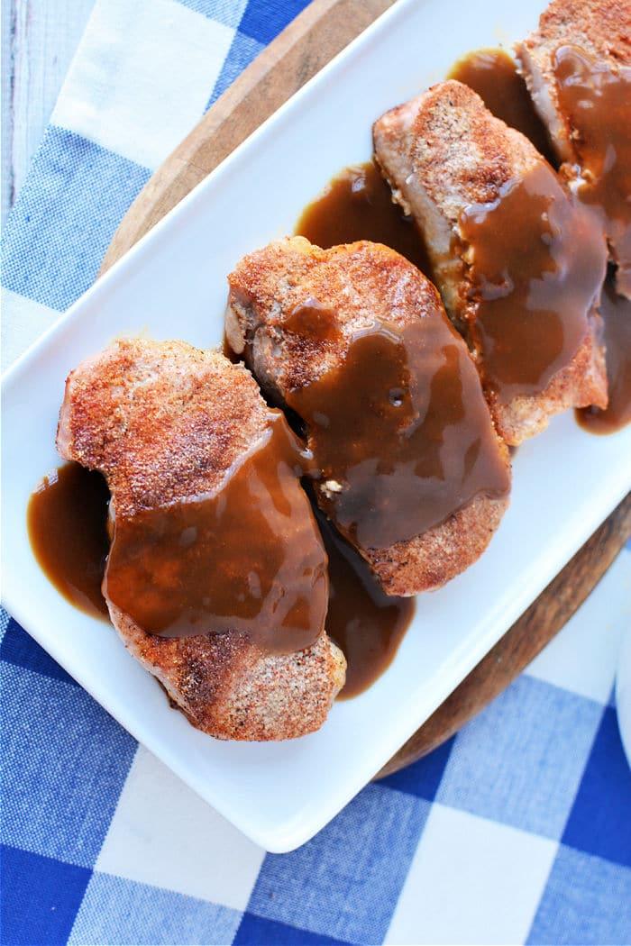 Four pieces of seasoned pork chops, expertly cooked from frozen, are topped with rich brown gravy and arranged on a rectangular white plate. The dish is beautifully set on a wooden board over a blue and white checkered tablecloth.