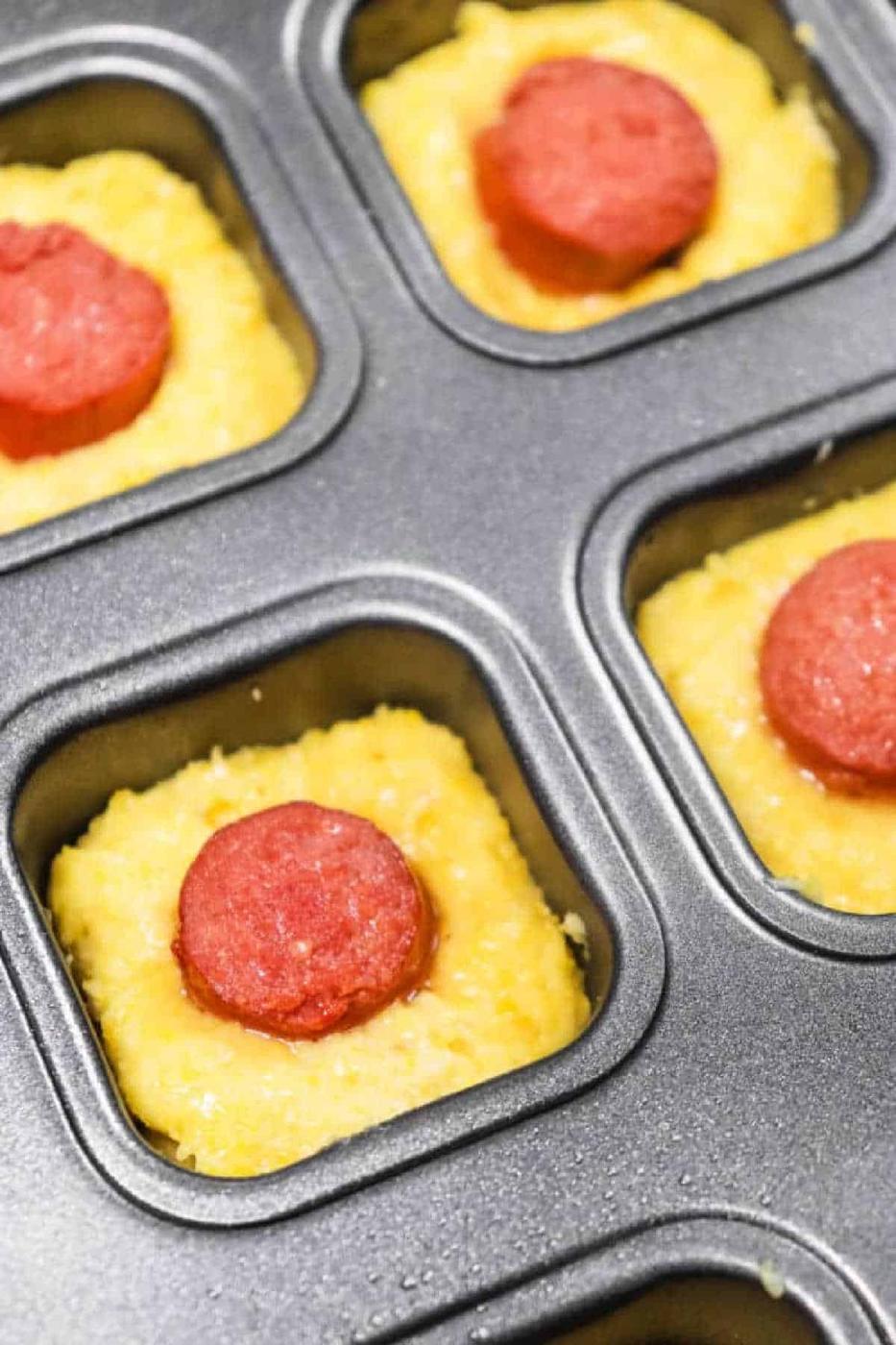 Unbaked Corn Dog Bites cornbread batter in a muffin tin, each topped with a slice of pepperoni.