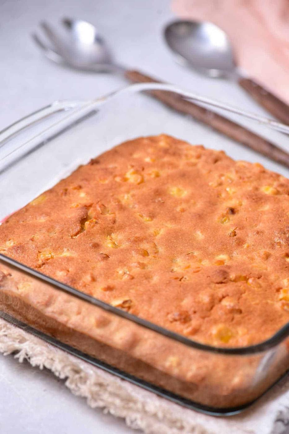 A square glass baking dish holds a freshly baked corn pudding with a golden-brown crust, reminiscent of homemade corn pudding without Jiffy mix. The dish is placed on a tan woven trivet, and two silver spoons are in the background on a light-colored surface.