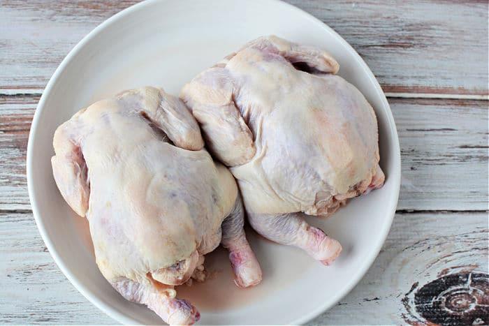 Two raw whole game hens rest side by side on a white plate atop a rustic wooden surface, evoking questions like how long to cook Cornish hen in an air fryer for the perfect meal.