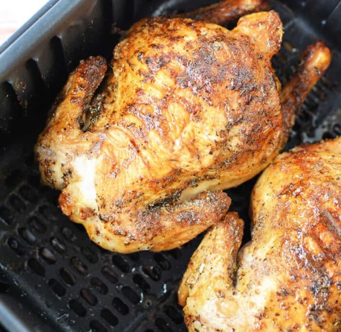 Two golden-brown roasted hens, reminiscent of slow cooker Cornish hens, rest in a black air fryer basket, seasoned with herbs and spices.
