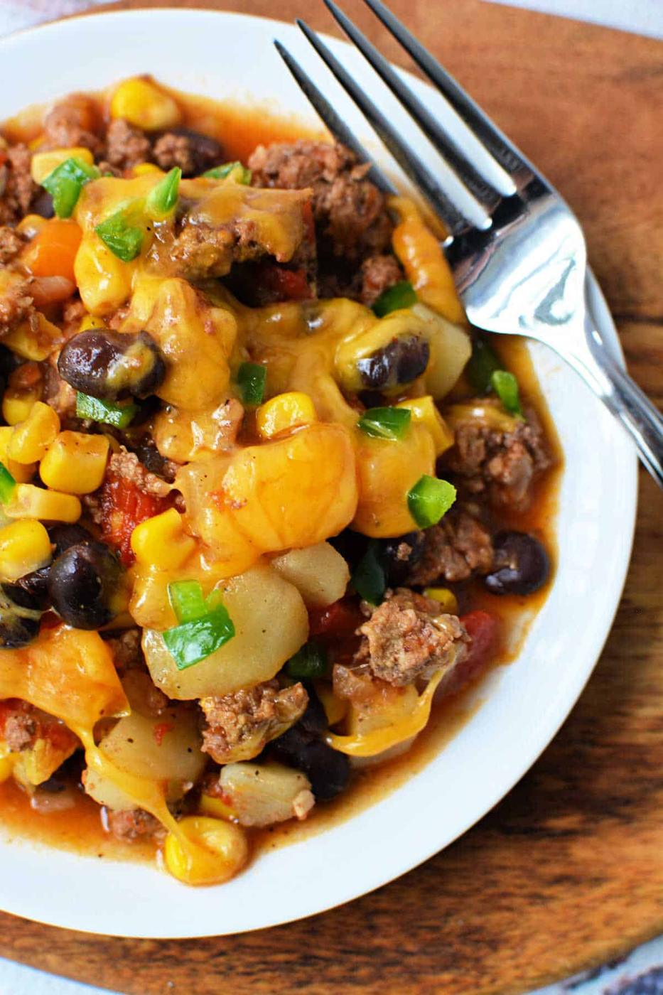 A plate of cheesy Cowboy Casserole with beef, black beans, corn, and potatoes sits next to a fork—perfect for fans of hearty Crockpot recipes.