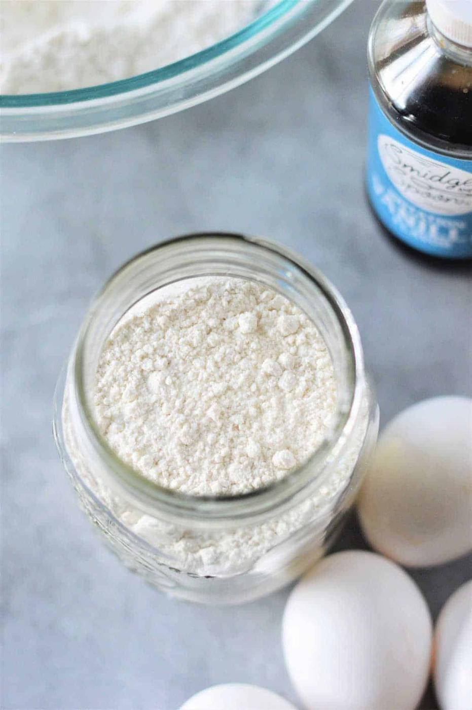 A jar filled with Cracker Barrel Pancake Mix, sits on the countertop next to eggs and a bottle of vanilla extract.