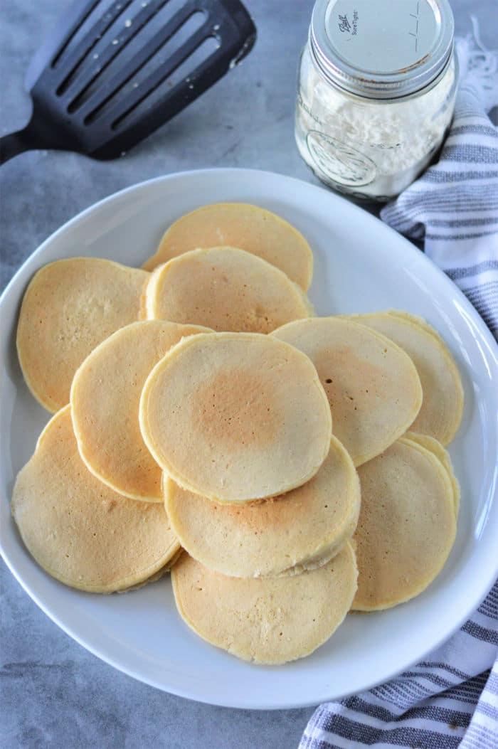 A white plate with a stack of uniformly sized, round pancakes, made from Cracker Barrel Pancake Mix, arranged neatly. A spatula and a jar with a white powder, likely flour, are placed in the background. A striped cloth napkin is visible on the right side of the plate.