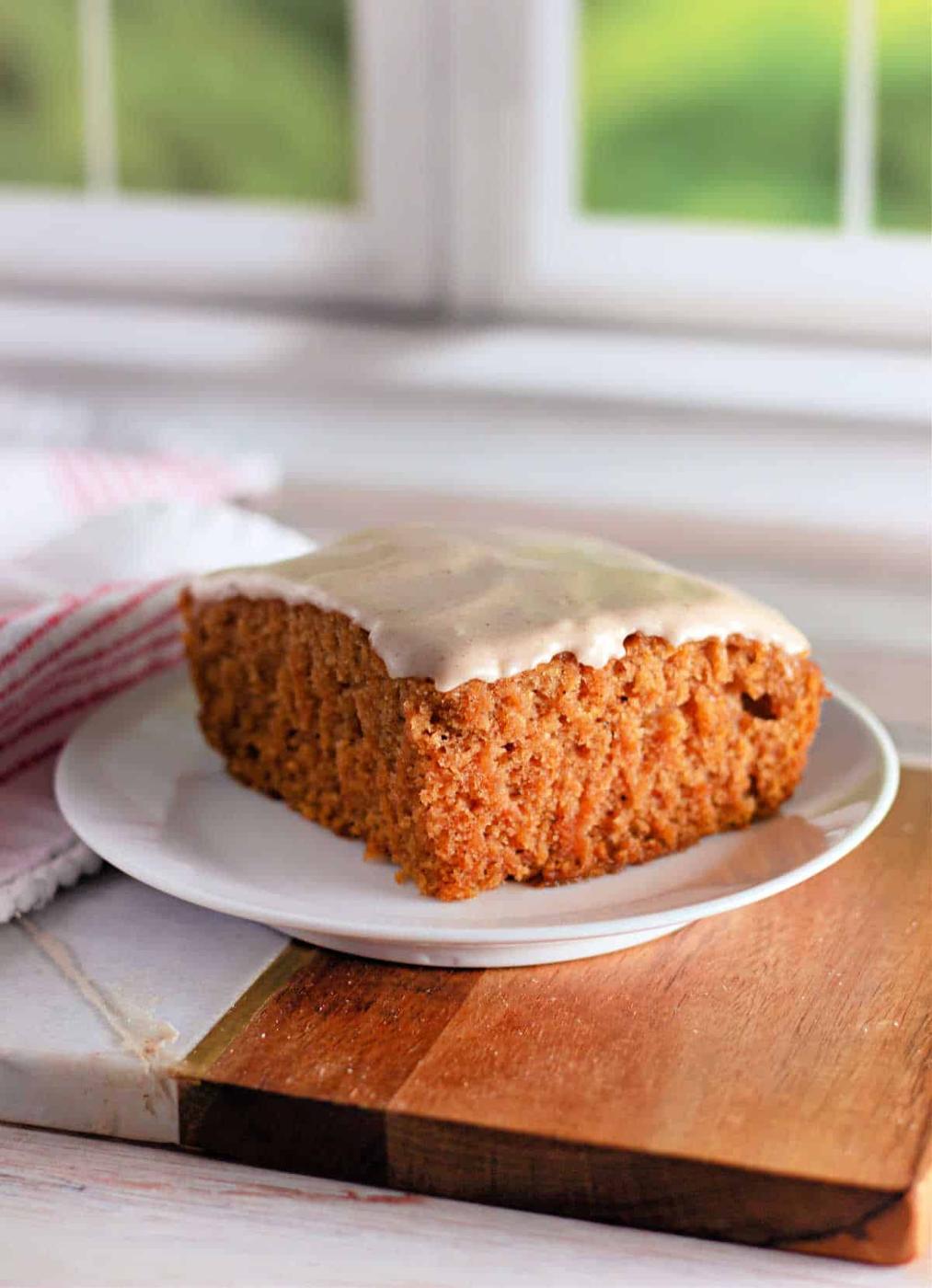 A slice of frosted Crazy Cake carrot cake sits on a white plate. The cake has a thick, soft texture and is topped with a light layer of white icing. The plate rests on a wooden board, with a white and red striped cloth partially visible in the background.