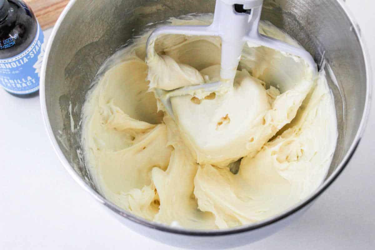 Creamy frosting in a mixing bowl with a paddle attachment, next to a bottle of vanilla extract, promises a rich flavor. Consider topping it with smooth cream cheese frosting without butter for an indulgent finish.