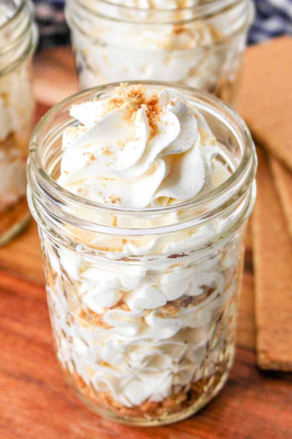 A jar filled with layers of cool whip frosting, crumbs, and a silky cream cheese frosting without butter, topped with more whipped cream and crumbs on a wooden surface.