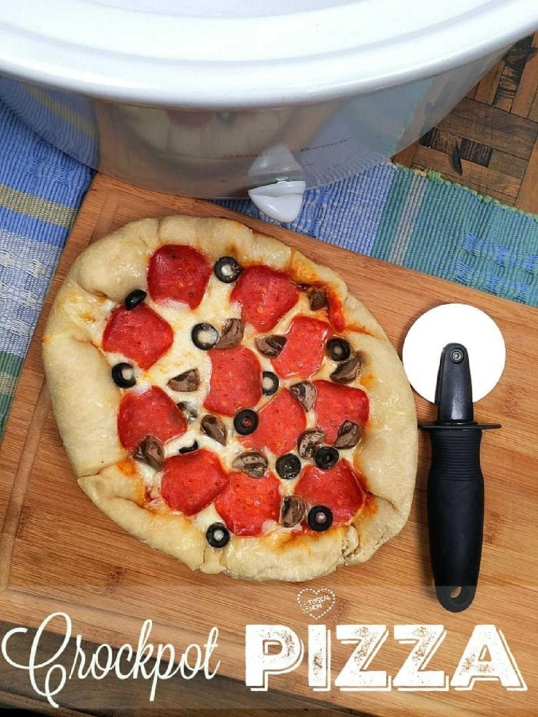 A homemade crock pot pizza with pepperoni, mushrooms, and olives rests on a cutting board next to a pizza cutter and crockpot.