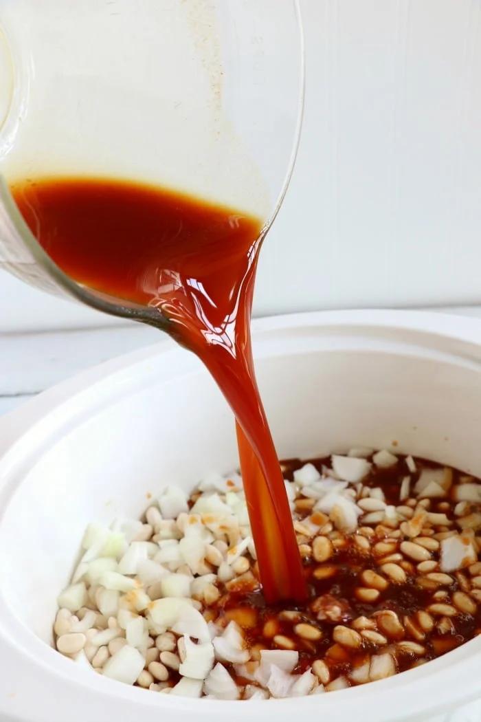 Pouring sauce into a slow cooker with chopped onions and white beans creates the perfect base for a homemade crockpot baked beans recipe.