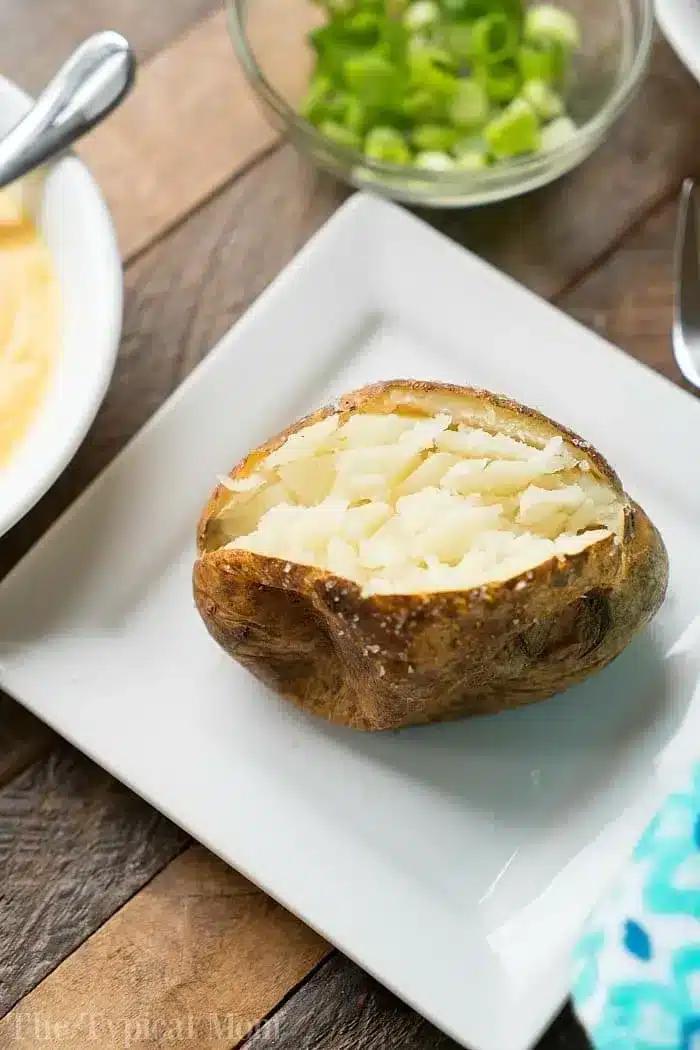 A baked potato with the top cut open, showing the fluffy interior, is placed on a white plate on a wooden table.