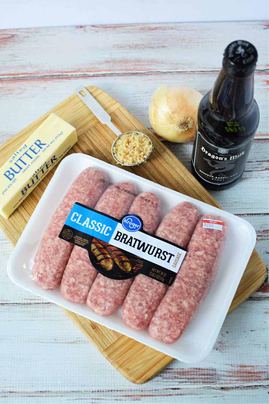 bratwurst, butter, chopped garlic, onion, and a beer bottle rest on a cutting board—perfect for crafting delicious Crockpot Brats.