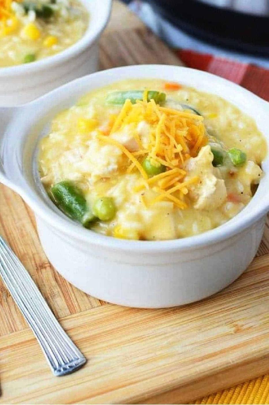 A bowl of creamy chicken and rice topped with shredded cheese on a wooden board, reminiscent of a comforting crockpot chicken casserole, sits invitingly beside a fork.