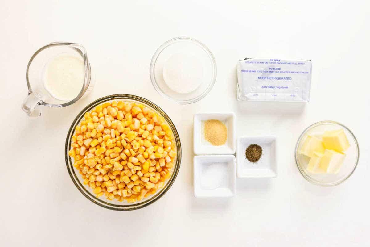 Bowls of corn, butter, seasonings, cream, and sugar on a white surface, ready for cooking the perfect Crockpot Creamed Corn.