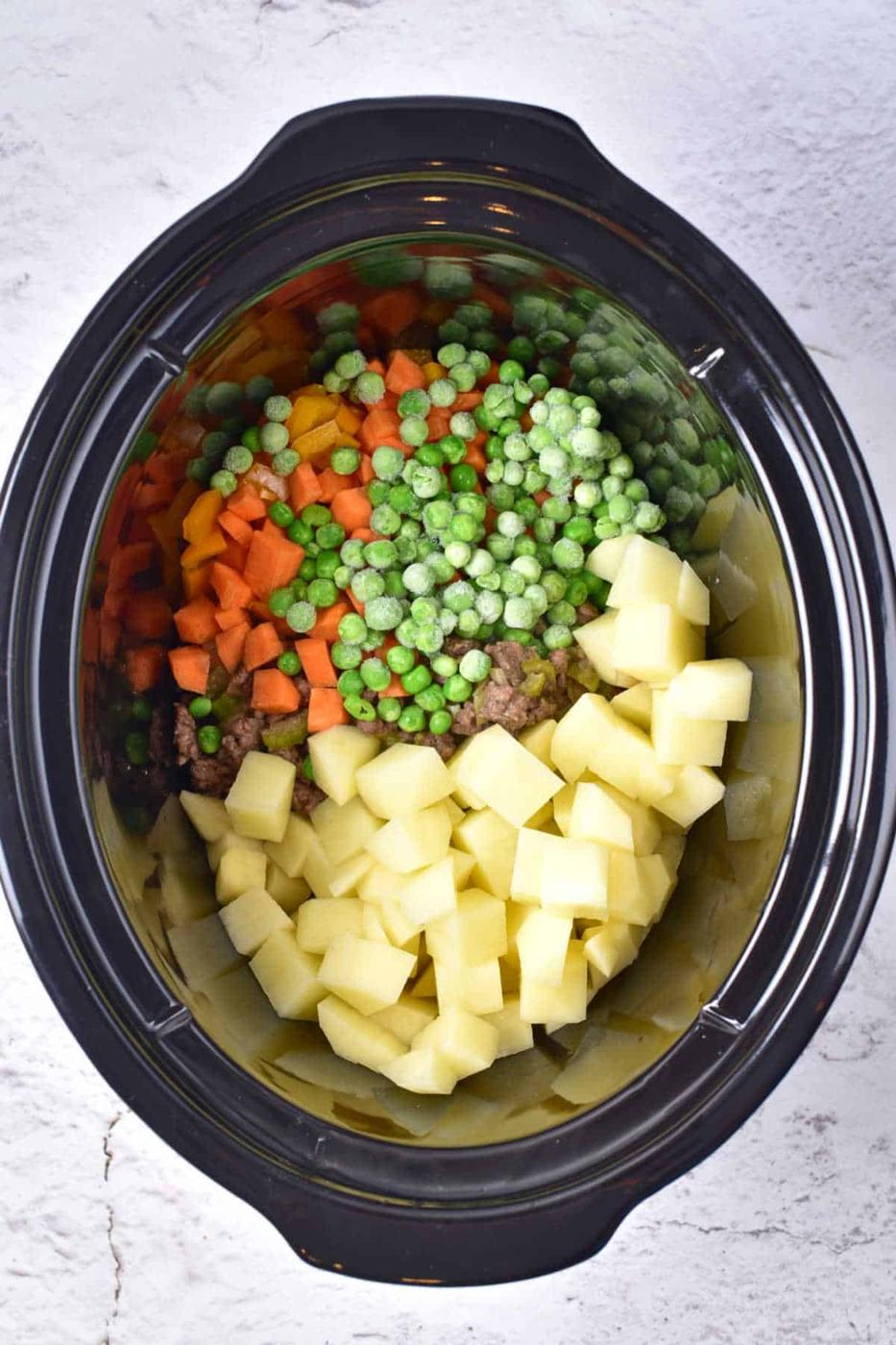A crockpot brimming with diced potatoes, carrots, peas, and sliced onions sits on a light gray surface, ready to transform into a creamy potato and hamburger soup.