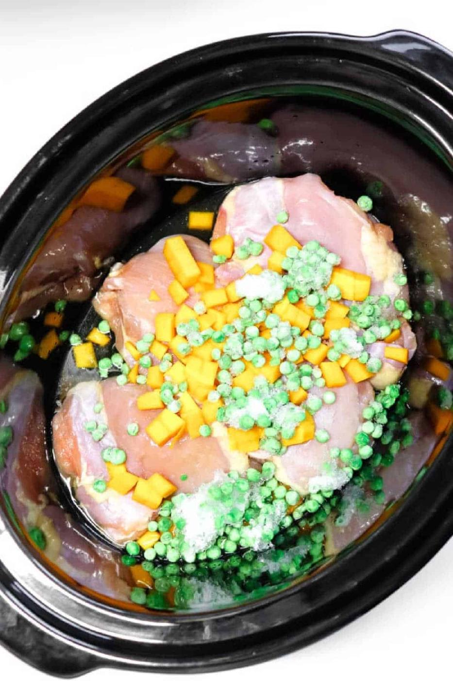 Raw chicken, frozen peas, and diced yellow vegetables in a black slow cooker, perfect for making wholesome Crockpot Dog Food.