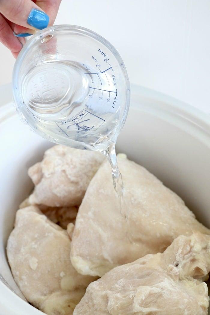 A hand holding a measuring cup pours water over frozen chicken breasts in a white crockpot, the uncooked chicken pale in color.