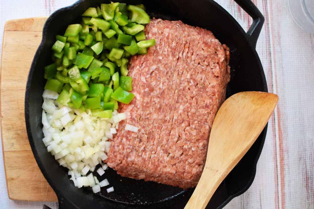 Ground meat, chopped green bell pepper, and chopped onion in a cast iron skillet with a wooden spatula—perfect for starting your Slow Cooker Lasagna Soup.