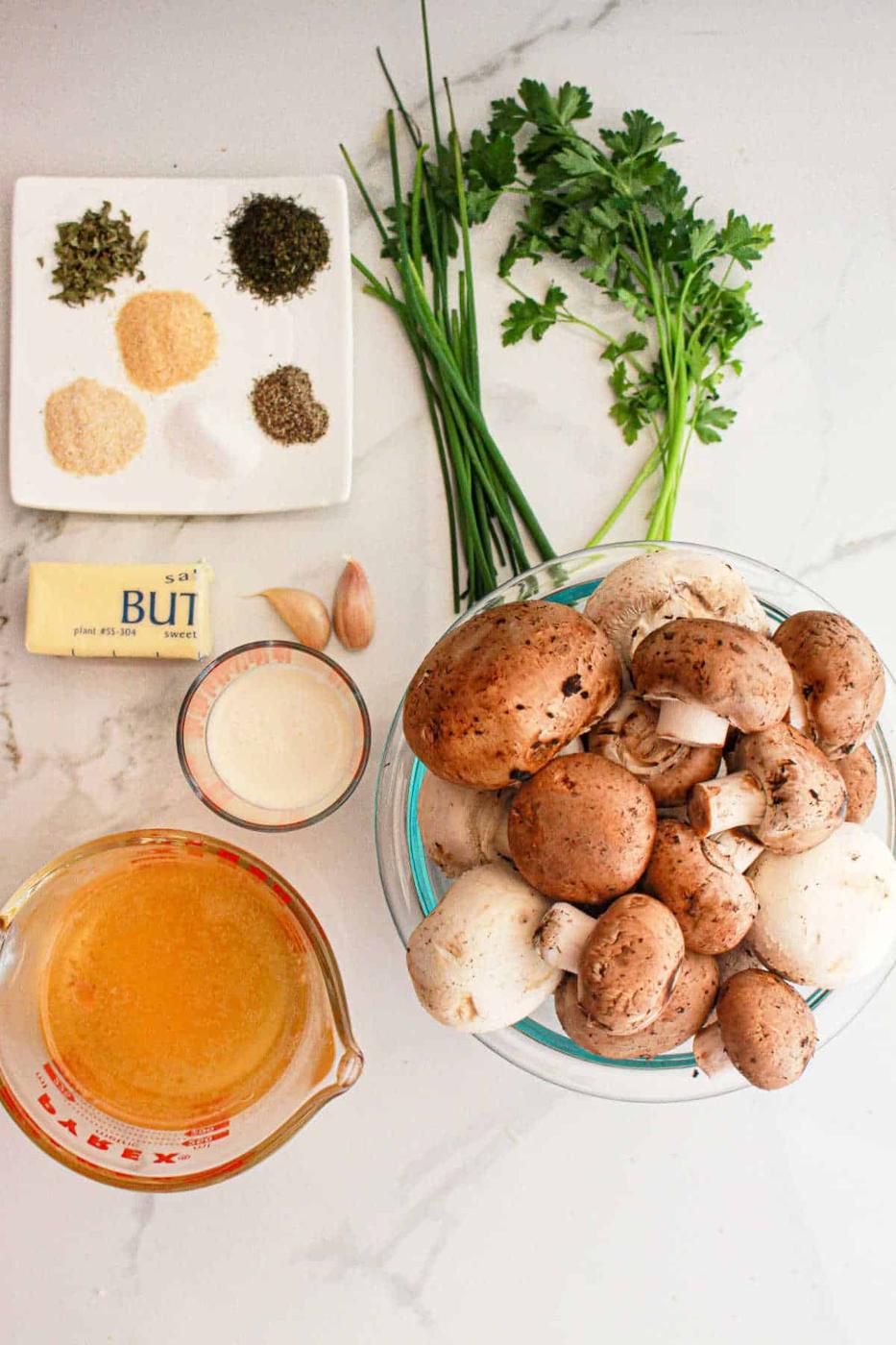 Ingredients for crockpot mushrooms: mushrooms, herbs, butter, garlic, cream, broth, parsley, and chives on a marble surface.