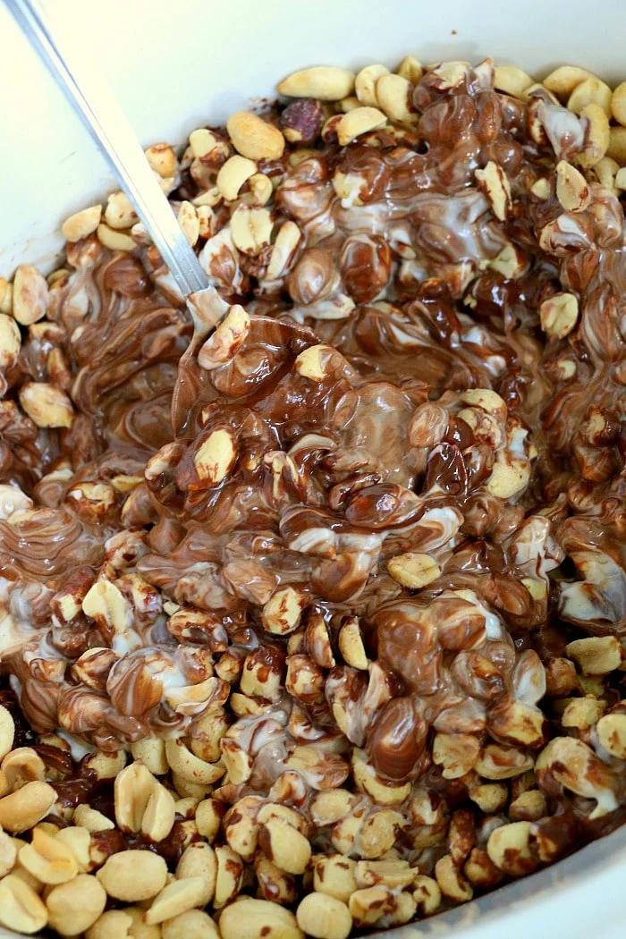 Peanuts mixed with melted chocolate and white cream, stirred together in a bowl with a metal spoon, create irresistible crock pot peanut clusters perfect for any sweet craving.
