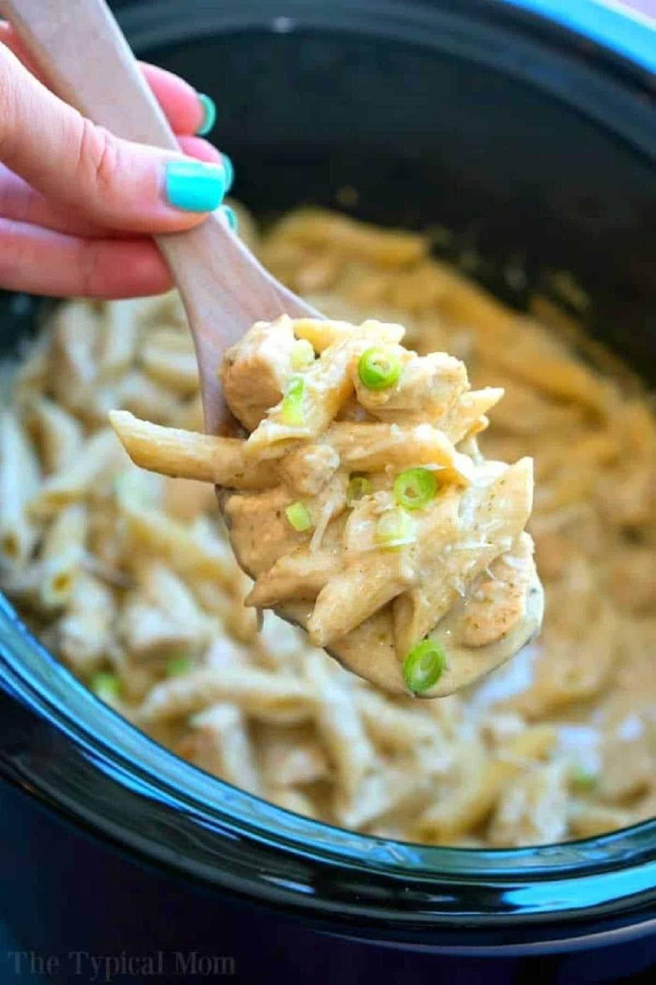 A wooden spoon holds creamy pasta with green onions over a crockpot pesto chicken alfredo casserole, brimming with rich flavors.