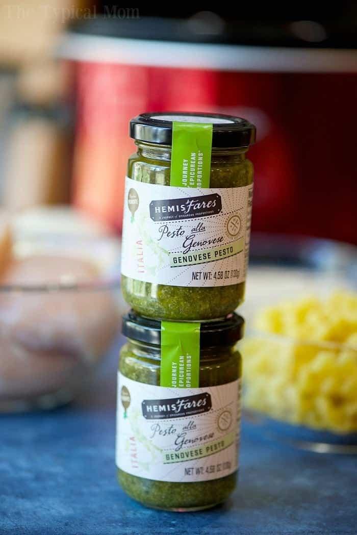 Two jars of HemisFares Genovese pesto stacked on a table with blurred ingredients in the background, perfect for adding bold flavor to your next crockpot pesto chicken alfredo casserole.