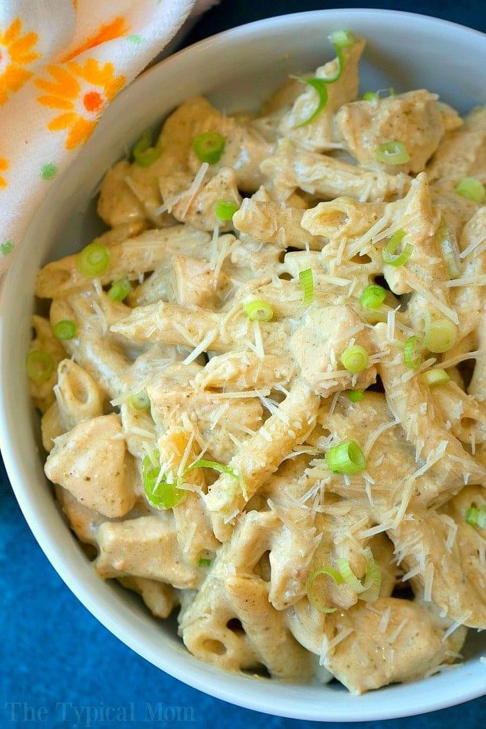 Bowl of creamy pasta with grated cheese and sliced green onions, next to a floral-patterned napkin—reminiscent of a hearty crockpot pesto chicken alfredo casserole.
