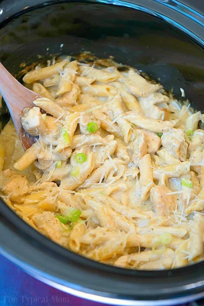 Creamy crockpot pesto chicken alfredo casserole with penne, cheese, and green onions, all blended together in a slow cooker with a wooden spoon.