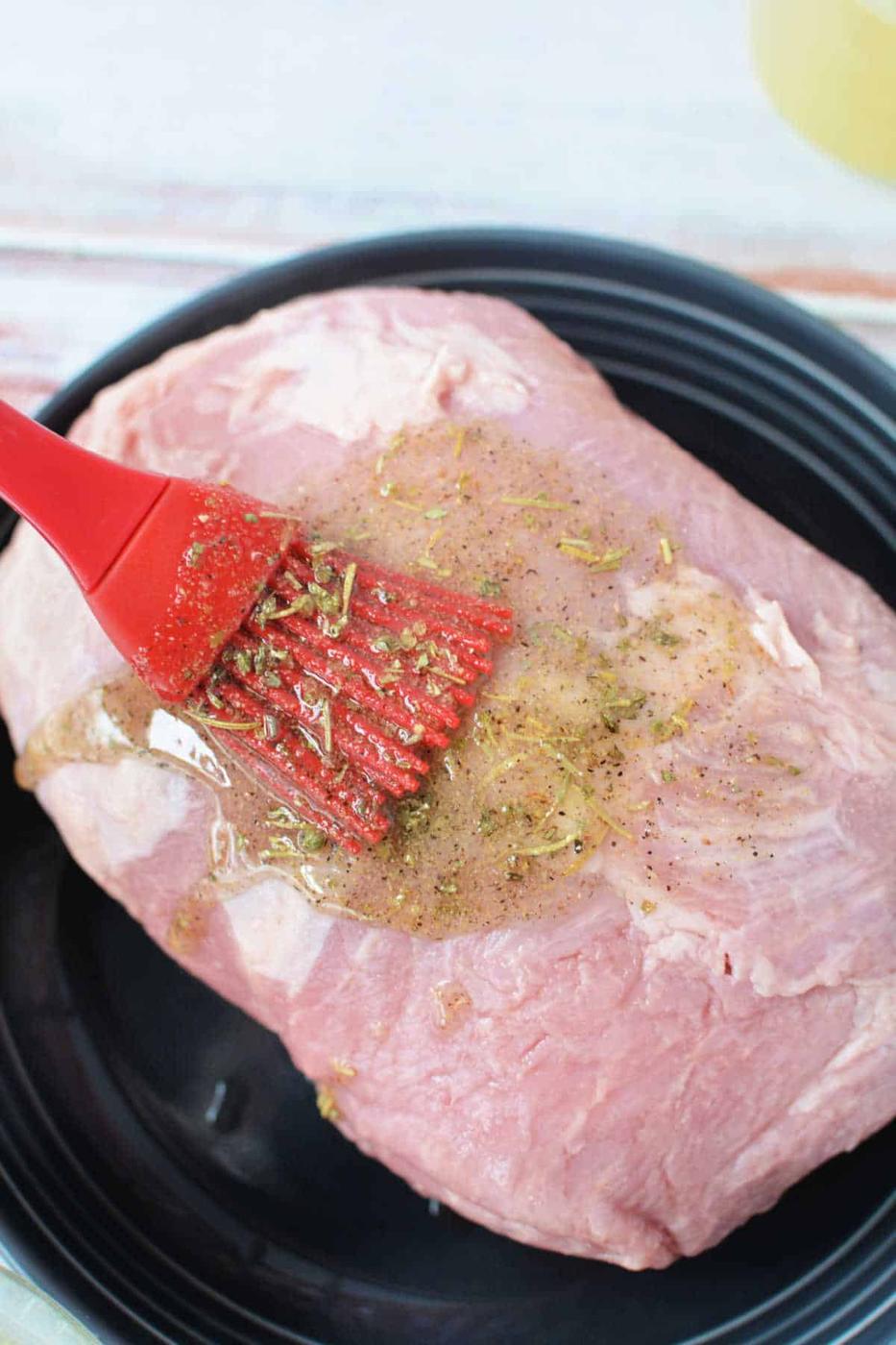 A Crockpot Porketta Roast is prepared as a delectable raw pork roast, with a flavorful marinade gently applied using a red brush on a sleek black plate.