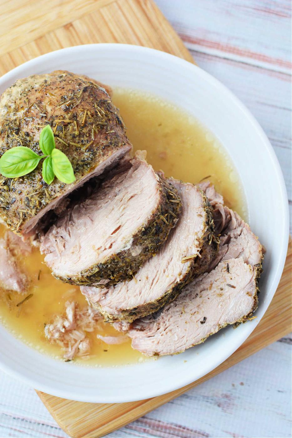 Enjoy a deliciously tender Crockpot Porketta Roast, perfectly herb-crusted and sliced, served with rich gravy on a white plate. Garnished with fresh basil for an added touch of flavor.