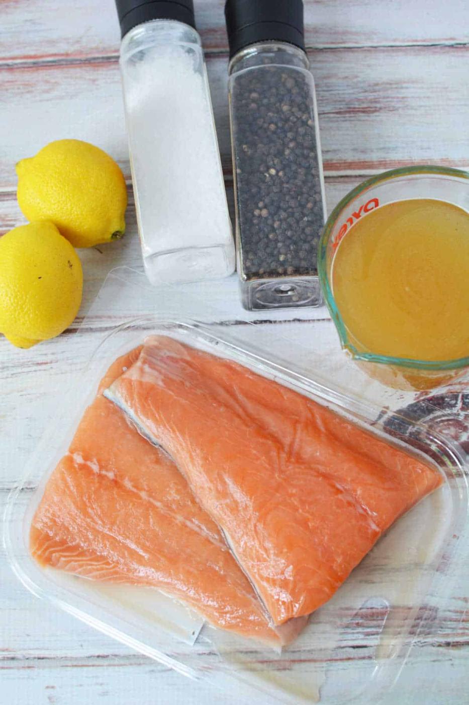 Raw salmon fillets in a plastic tray with lemons, salt, pepper, and a measuring cup of broth—perfect ingredients for easy Crockpot Salmon on your table.