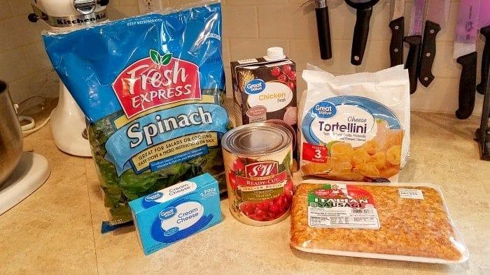 Ingredients for crockpot tortellini soup on a counter: spinach, cream cheese, diced tomatoes, sausage, tortellini, and chicken broth.