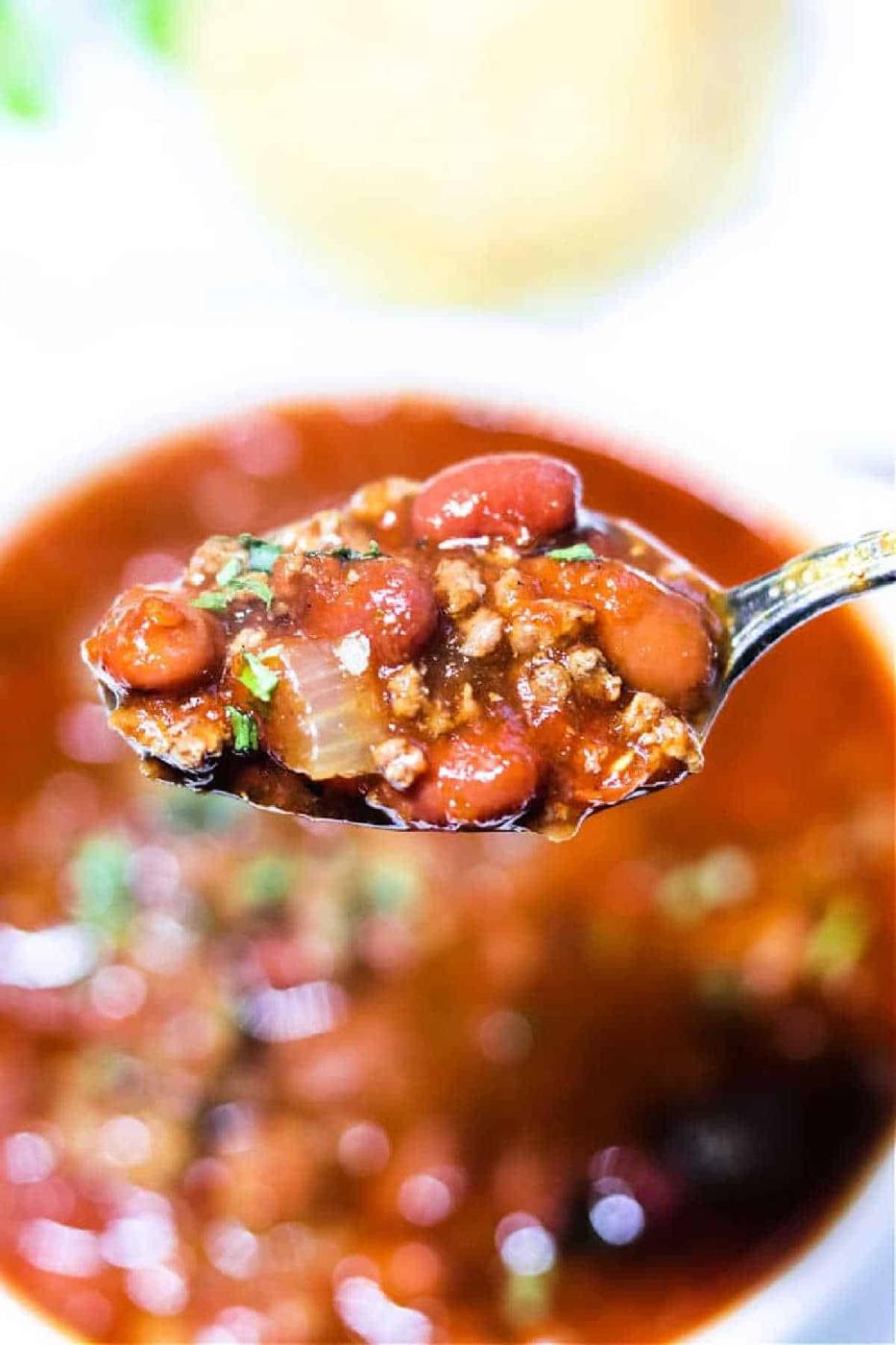 A spoonful of venison chili with beans, ground meat, and onions held above a bowl of hearty chili.