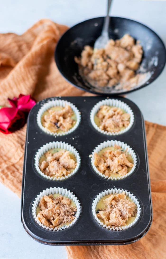 A muffin tray with six unbaked blueberry muffins with frozen blueberries sits next to a bowl of crumb topping and a red rose on a brown cloth.
