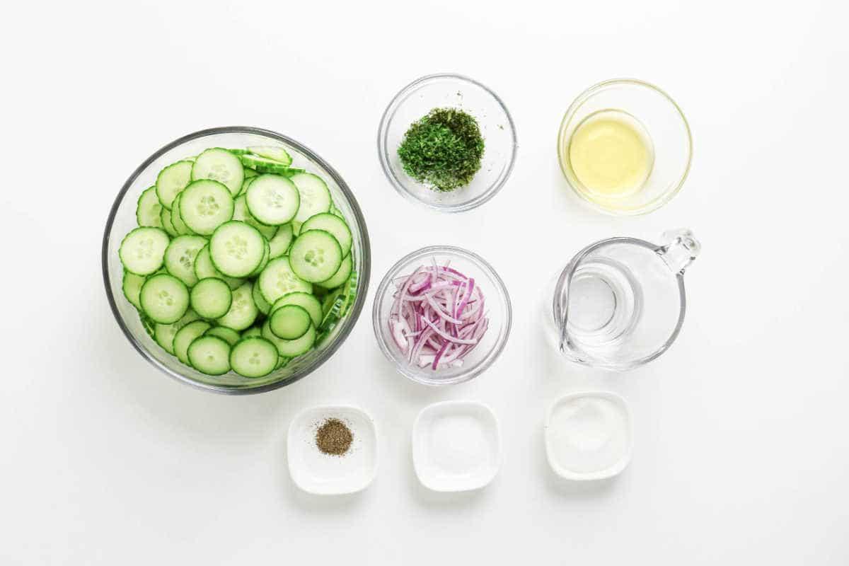 Sliced cucumber, red onion, fresh dill, vinegar, water, salt, pepper, and sugar in bowls on a white background—perfect ingredients for a refreshing cucumber salad.