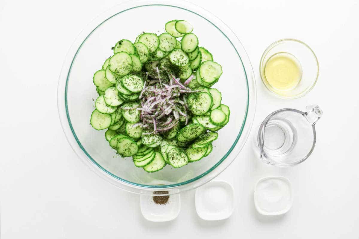 Glass bowl with sliced cucumbers, onions, and fresh dill, creating a crisp cucumber salad, surrounded by vinegar, water, salt, sugar, and pepper.