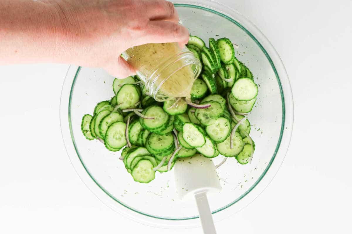 A hand pours dressing over sliced cucumber and onion in a glass bowl with a white spatula, creating a fresh salad perfect for adding dill.
