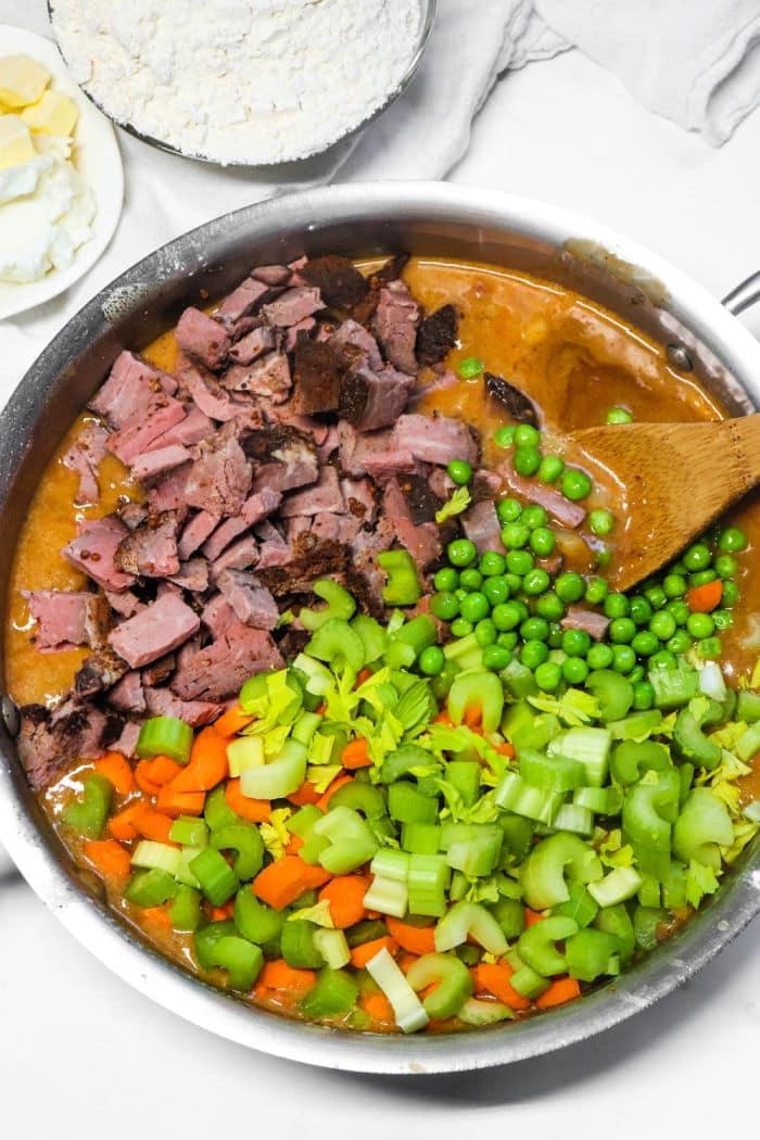 A skillet with diced meat, peas, chopped celery, carrots, and creamy sauce being stirred with a wooden spoon—the perfect filling for a comforting Double Crust Pot Pie.