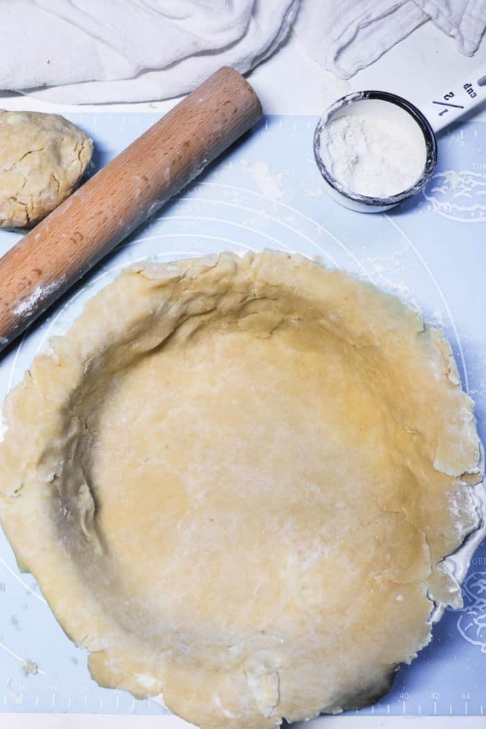 Unbaked pie crust in a pan, with a rolling pin, dough ball, and cup of flour nearby on a blue surface—perfect foundations for a homemade Double Crust Pot Pie.