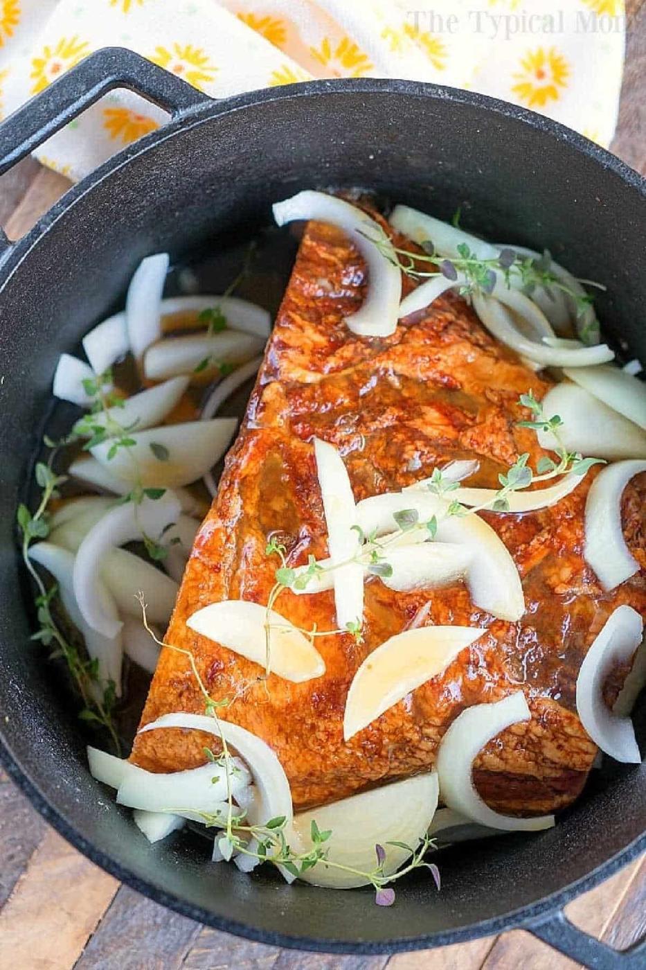 Seasoned dutch oven ham in a black pot, topped with sliced onions and fresh herbs, ready to be cooked.