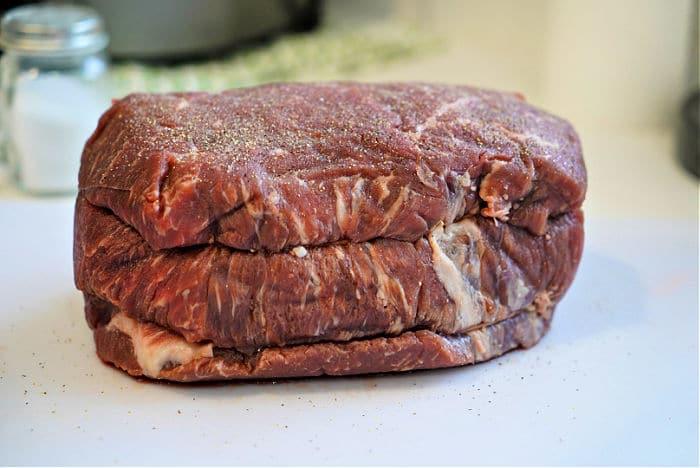 A raw beef roast sits on a white cutting board, lightly seasoned with salt and pepper, ready to become a tender Dutch oven roast. A salt shaker and a green and white cloth are blurred in the background.