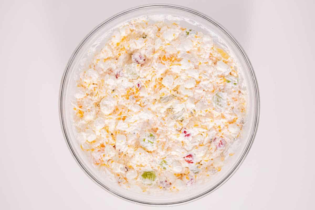 A glass bowl filled with a creamy salad mixture containing marshmallows, fruit, and shredded cheese—an easy ambrosia recipe perfect for any occasion.
