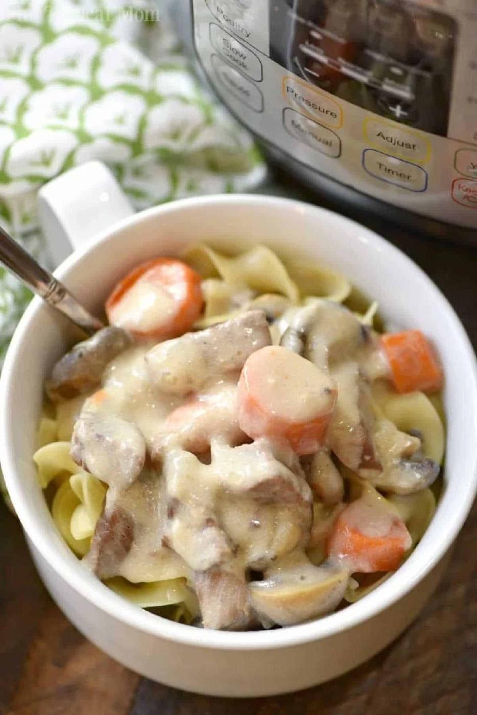 A bowl of Instant Pot beef stroganoff with stew meat, carrots, and mushrooms served over noodles sits next to the pressure cooker.
