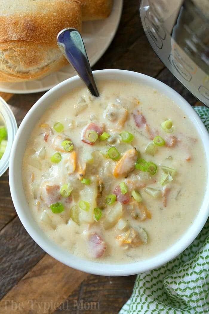 A bowl of creamy clam chowder, made effortlessly as easy instant pot clam chowder, is garnished with chopped green onions on a wooden table. Beside it sits a loaf of bread and the partially visible pressure cooker. A green and white cloth peeks out next to the bowl.