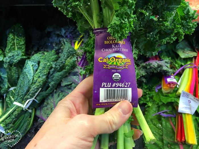 A hand holds a bunch of organic kale with a purple label in a grocery store, offering inspiration for easy kale recipes.