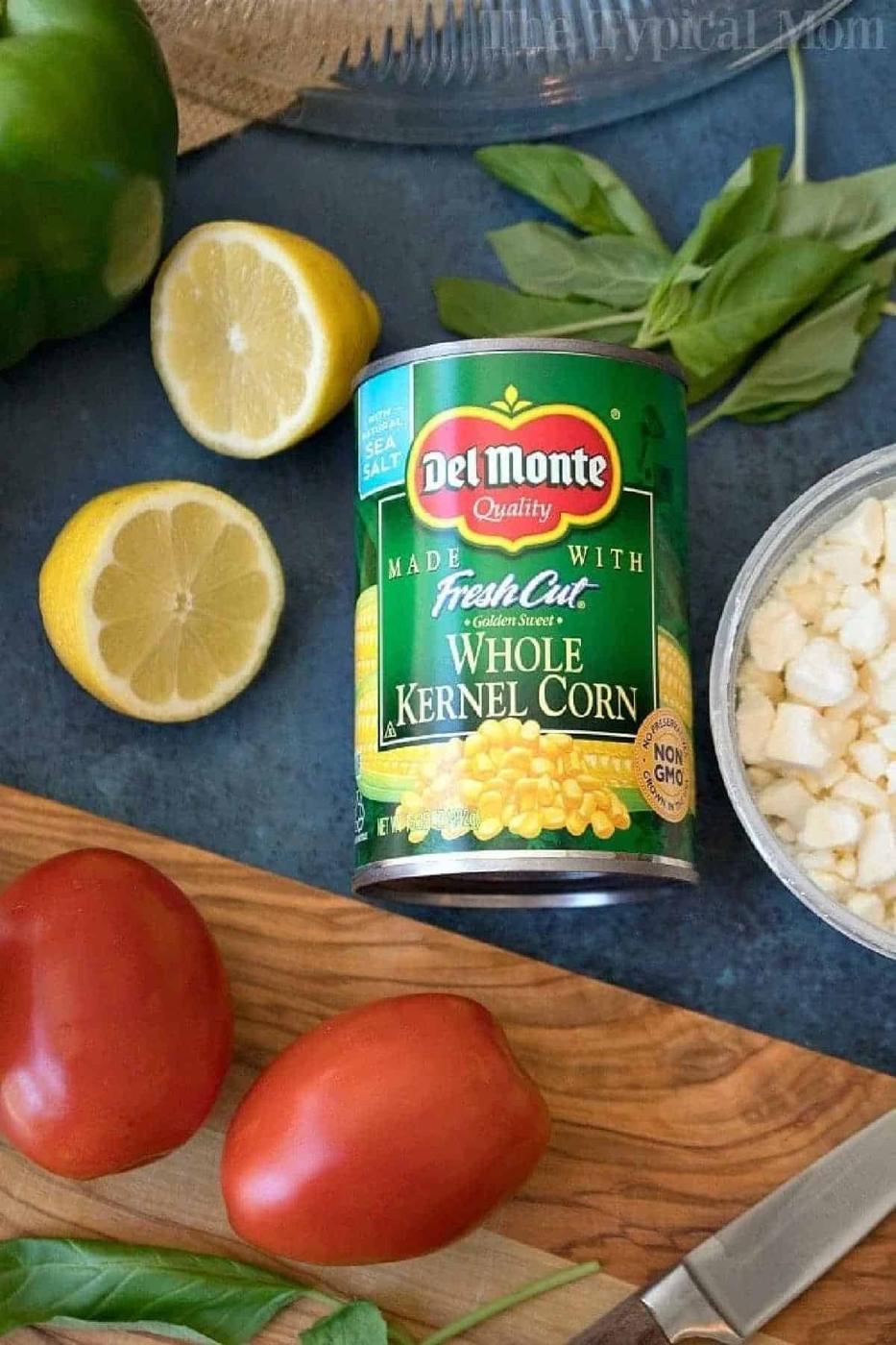 A can of Del Monte whole kernel corn surrounded by tomatoes, lemon, basil, and chopped onions—perfect ingredients for a fresh Jiffy cornbread salad.