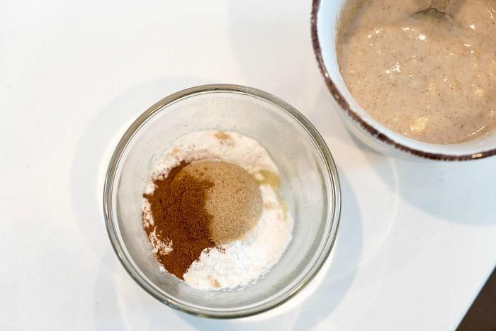 Glass bowl with flour, sugar, and cinnamon sits beside a mug containing a mixed, beige batter with a spoon—perfect for an easy mug cake recipe.