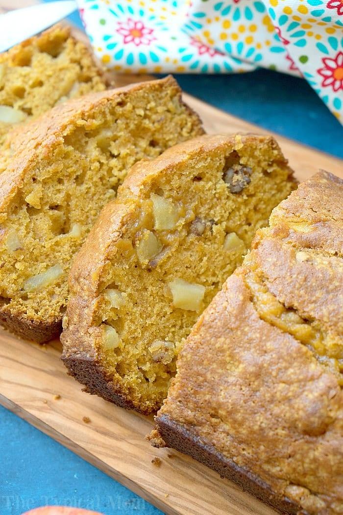 Slices of easy pumpkin apple bread with walnuts rest on a wooden board, accompanied by a floral cloth in the background.