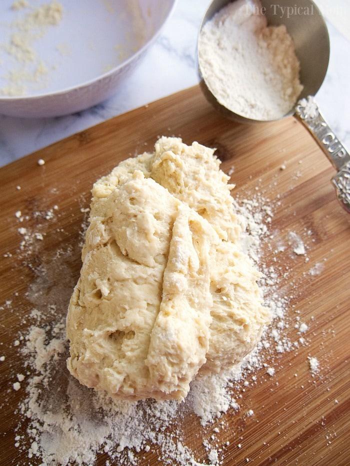Dough on a floured wooden board with a metal measuring cup of flour nearby, ready to start your easy roll recipe.