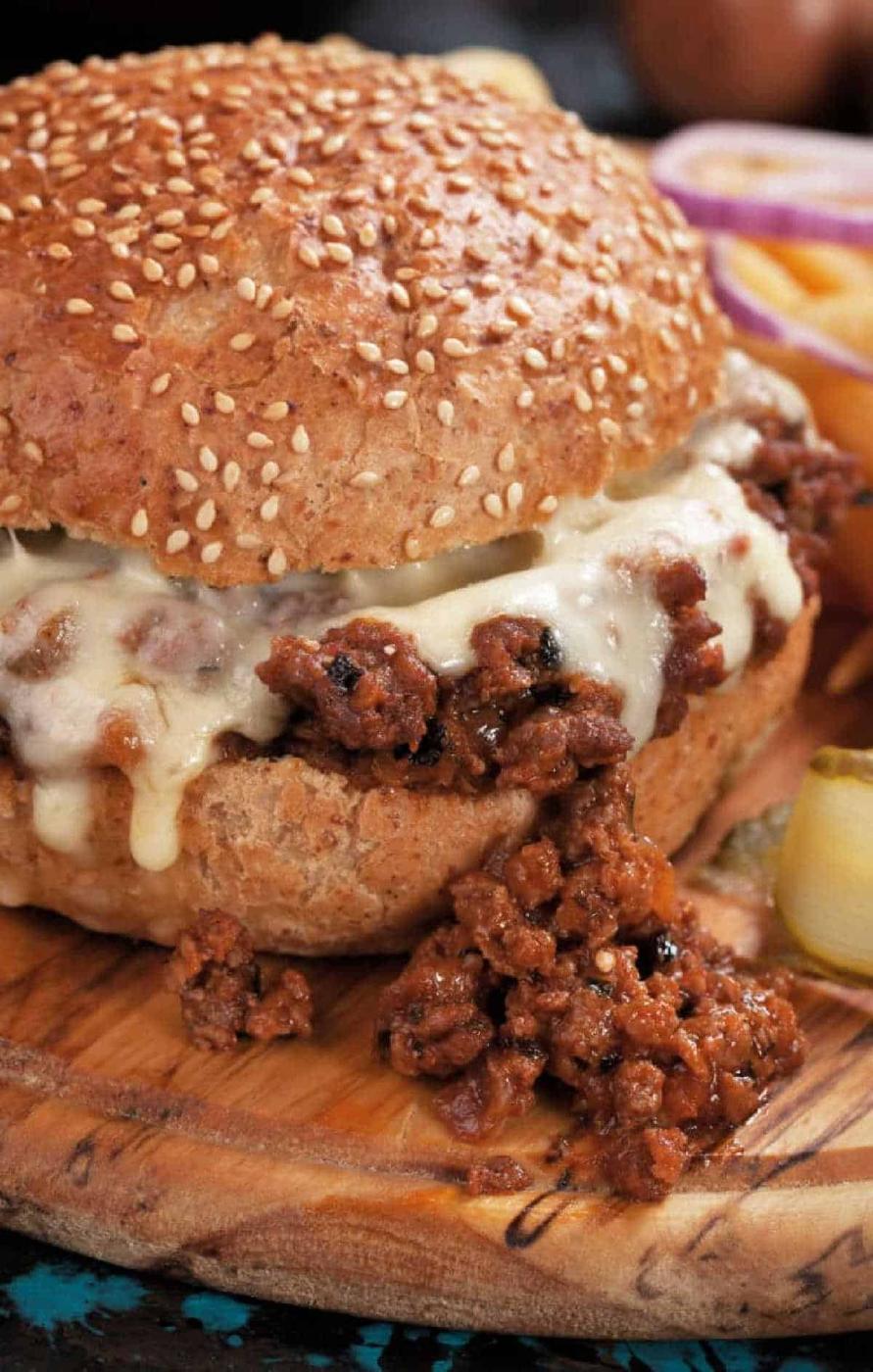 Close-up of a sloppy joe sandwich made with an easy sloppy joe recipe 3 ingredients, topped with melted cheese on a sesame bun, served on a wooden board.