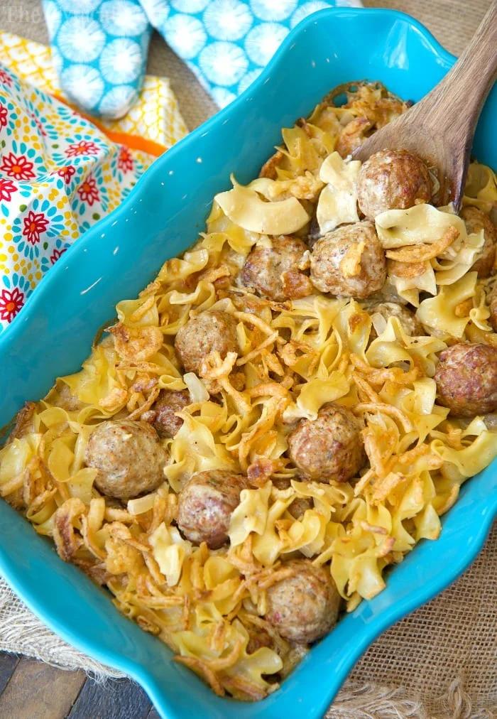 A blue baking dish brimming with pasta and an easy Swedish meatball casserole, topped with a creamy sauce. Baked to perfection, it rests beside a wooden spoon. A colorful patterned cloth adds a touch of warmth in the background.